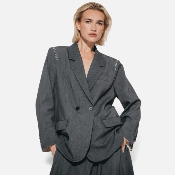 1. Frau trägt grauen Oversize Blazer mit tiefem Revers, Frontalansicht 2. Grauer zweireihiger Oversize Blazer, Flatlay-Ansicht 3. Rückenansicht des grauen Oversize Blazers mit Schulterdetail 4. Detailansicht Ärmelknöpfe und Taschen des Oversize Blazers 5. Nahaufnahme Revers mit Kontrastpaspel und Steppnaht-Detail 6. Model im kompletten Look mit grauem Oversize Blazer und Midirock